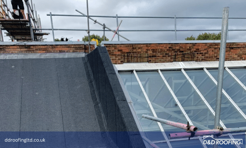 Close-up of a newly installed flat felt roof junction with a pitched glazed roof section, showing precise detailing and clean lines. Scaffolding surrounds the brick building exterior.