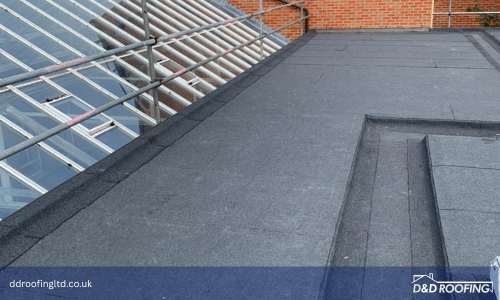 Wide view of the completed flat roof area, with clean felt seams and precise perimeter edging next to a sloped glass structure and red-brick building.